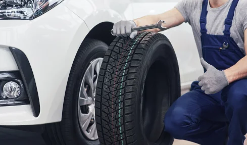 A man is changing a tire, holding a tire next to a car in a roadside setting.