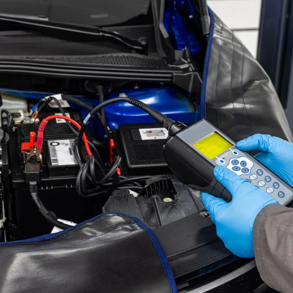 A person in blue gloves uses a handheld device to check a car battery during diagnostics.