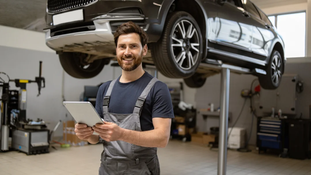 A mechanic in an auto repair shop examines a tablet while surrounded by tools and vehicle parts.